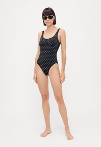 Black swimsuit with white polka dots, high-cut leg design, wide straps, and smooth texture, displayed on a standing figure wearing sunglasses.