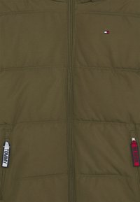 Oliven grøn pufferjakke med lynlåsåbning, der har to sidelommer med kontrasterende trækremme og et lille logo på brystet.