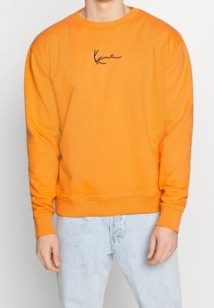 Man draagt een feloranje sweatshirt met lange mouwen en een zwart scriptlogo op de borst, gecombineerd met lichtblauwe spijkerbroek, staand tegen een effen achtergrond.