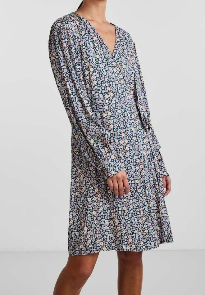 Femme portant une robe fleurie à manches longues, longueur genou, avec des fleurs roses, blanches et bleues sur un fond sombre.
