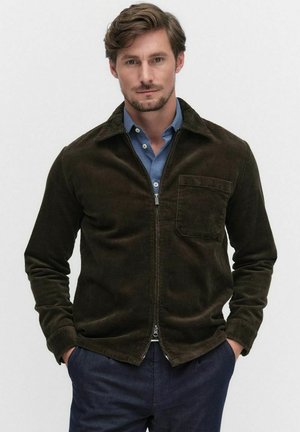 Brown corduroy jacket with a zipper, pointed collar, and front pocket. Worn over a blue shirt, model stands with hands in pockets.