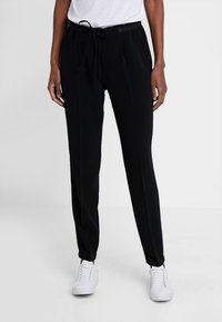Pantalon de jogging noir avec taille à cordon, plis latéraux et coupe décontractée. Fabriqué en matériau doux, assorti à des baskets blanches.