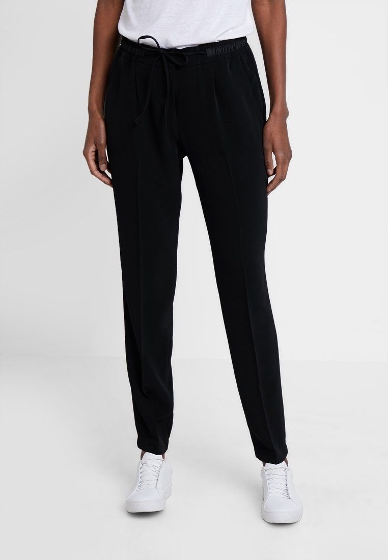 Pantalon de jogging noir avec taille à cordon, plis latéraux et coupe décontractée. Fabriqué en matériau doux, assorti à des baskets blanches.