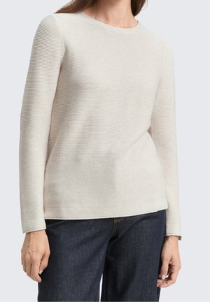 Femme portant un pull côtelé à manches longues couleur crème associé à un jean denim bleu foncé, debout devant un fond clair uni.