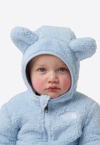 Combinezon de zăpadă din fleece albastru deschis cu urechi de urs pe glugă. Textură moale, fermoar complet și detaliu cu logo pe piept.