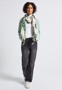 Kvinna med lockigt hår som bär en mintgrön jacka, vit skjorta, svarta jeans, vita sneakers och en mönstrad beige och mörkgrön scarf, går framåt.