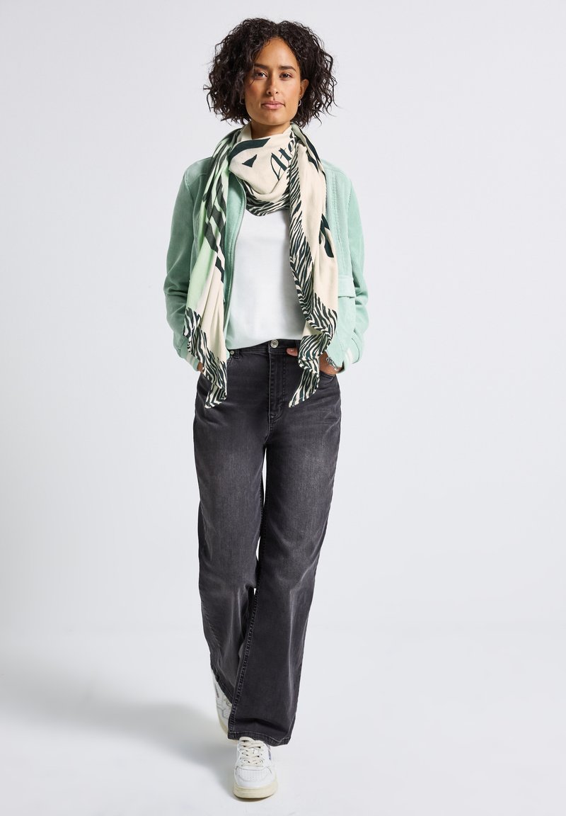 Kvinna med lockigt hår som bär en mintgrön jacka, vit skjorta, svarta jeans, vita sneakers och en mönstrad beige och mörkgrön scarf, går framåt.