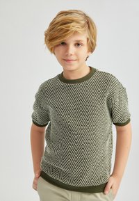 Suéter corto de mangas cortas en verde y blanco con un patrón de pata de gallo y cuello acanalado, combinado con pantalones de color claro.