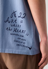 Tricou din bumbac de culoare bleu deschis, cu text bleu închis și o mică grafică. Textul include "Arta e acolo unde e inima" și detalii despre marcă.