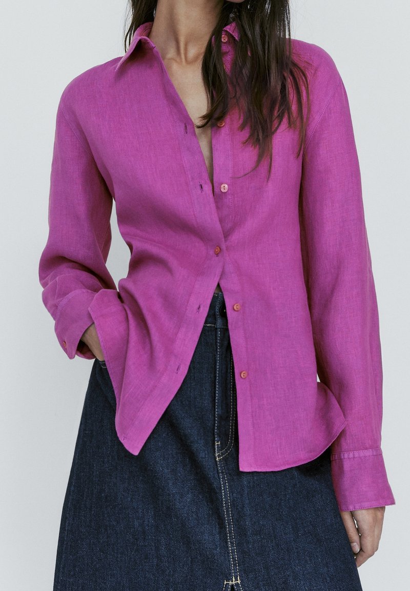Chemise en lin fuchsia avec un col et un devant à boutons, dotée de manches longues et d'une coupe décontractée. Portée avec une jupe en denim foncé.