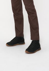 Czarne wysokie trampki z zamszu z gumową podeszwą, z sznurowanym zapięciem i brązową podeszwą. Połączone z brązowymi spodniami.