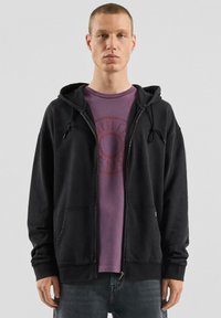 Czarna bluza z kapturem zapinana na zamek, z sznurkami i przednimi kieszeniami, noszona na fioletowej koszulce z okrągłym logo. Luźny krój i miękka faktura.