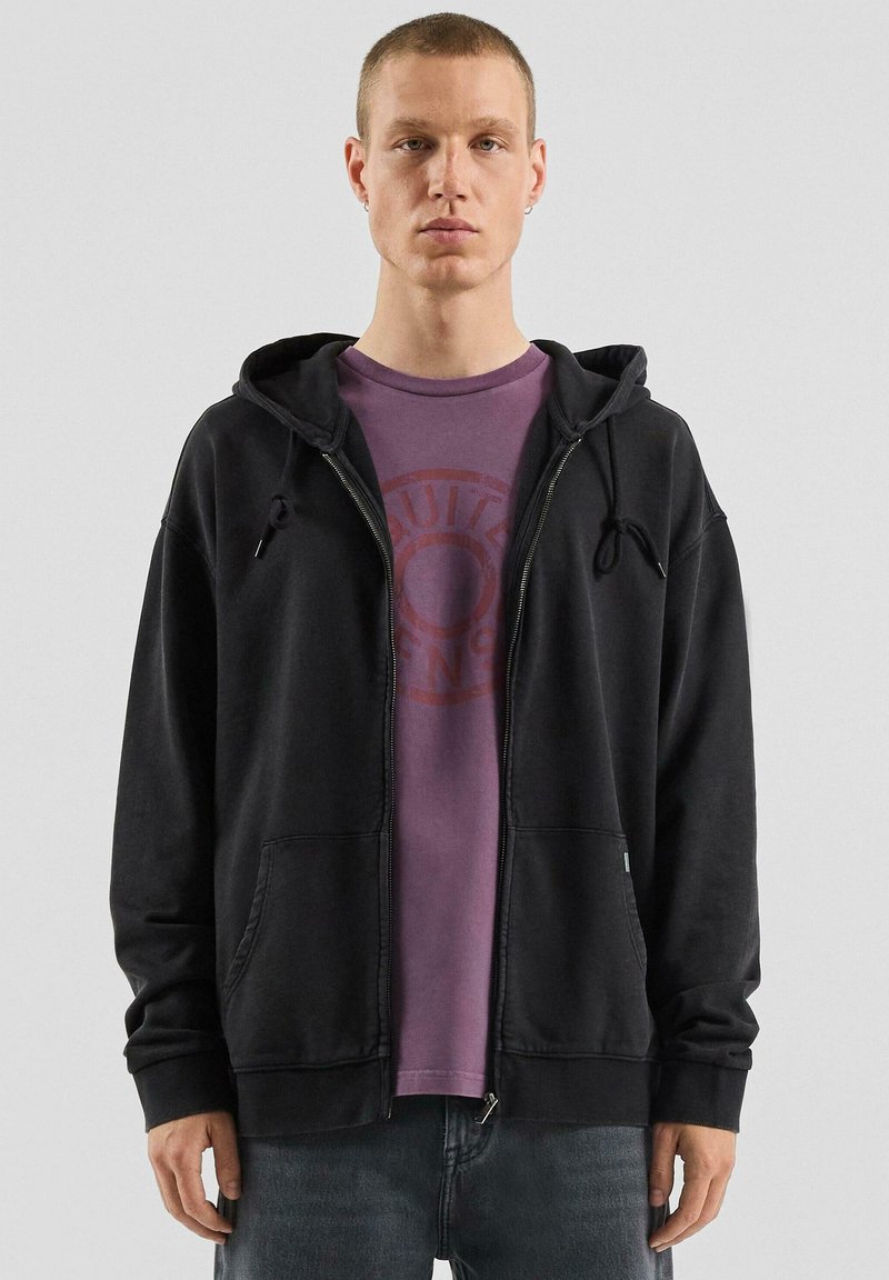 Czarna bluza z kapturem zapinana na zamek, z sznurkami i przednimi kieszeniami, noszona na fioletowej koszulce z okrągłym logo. Luźny krój i miękka faktura.