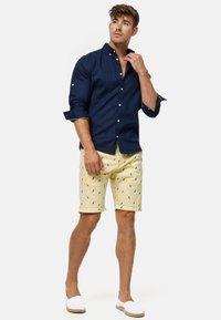 Chemise bleu marine à boutons avec les manches retroussées, assortie à un short jaune orné d'un petit motif d'oiseau et des chaussures blanches à enfiler.
