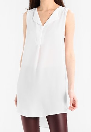Femme portant une tunique blanche sans manches avec un col en V et un ourlet asymétrique, associée à un pantalon ajusté marron foncé.
