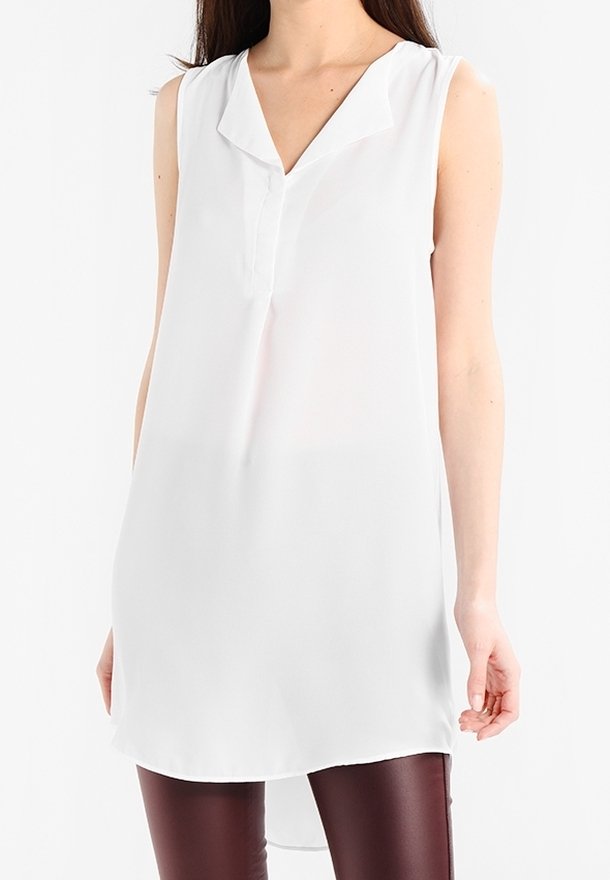 Femme portant une tunique blanche sans manches avec un col en V et un ourlet asymétrique, associée à un pantalon ajusté marron foncé.