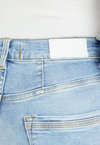 Jeans de mezclilla de color azul claro con una textura suave, que cuentan con una etiqueta blanca en la parte trasera y costuras amarillas en contraste a lo largo de las costuras.