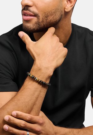 Mann mit kurzem Bart, der ein schwarzes Hemd trägt, zeigt ein Perlenarmband und einen silbernen Ring an der linken Hand vor schlichtem Hintergrund.