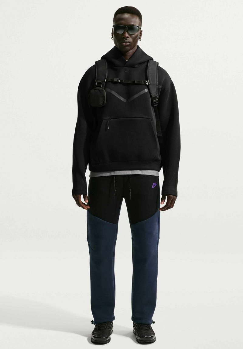 Sudadera negra con capucha y bolsillo canguro, combinada con pantalones a bloques de color en negro y azul marino, complementada con una pequeña bolsa y zapatillas.