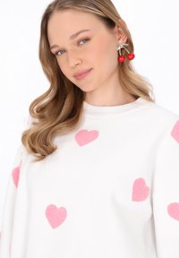 Hanorac alb cu modele de inimi din fleece roz; model purtând cercei în formă de cireșe roșii; textură netedă, design cu guler rotund.
