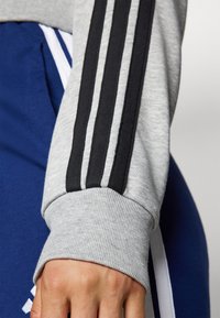 Grijze sweatshirtmouw met zwart drie-strepen ontwerp; geribbelde manchette zichtbaar; blauwe broek op de achtergrond met witte accenten.