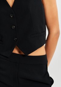 Czarna krótka kamizelka z dekoltem w serek, z pięcioma guzikami, dwiema bocznymi kieszeniami i dopasowanym krojem. Połączona z pasującymi czarnymi spodniami.
