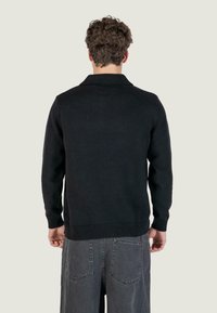 Maglione nero lavorato a maglia con colletto, polsini e orlo a coste, indossato su pantaloni grigi. La trama sembra morbida e accogliente.