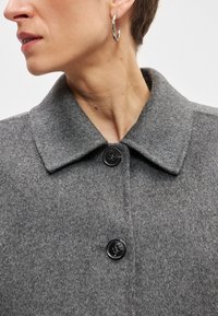 Γκρι παλτό από μαλλί με φαρδιά γιακά, με μαύρα κουμπιά και λεία υφή. Κοντινό πλάνο στο ντεκολτέ και ορατά σκουλαρίκια.