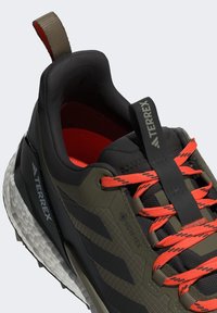 Black and olive green athletic shoe with textured upper, orange laces, and a white, cushioned sole, featuring a GORE-TEX label.