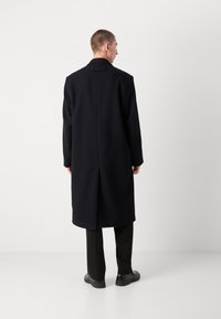 Abrigo negro de lana con corte recto, mangas largas y abertura trasera; presenta un cuello y un diseño minimalista. Combinado con pantalones negros.