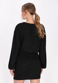 Schwarzes Kleid mit langen Ärmeln, gefertigt aus strukturiertem Stoff, der farbenfrohe Glitzerpartikel enthält. Taillierter Schnitt mit leicht ausladendem Saum.