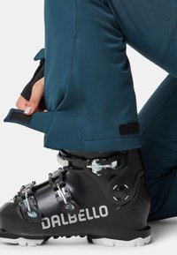 Dalbello-Skischuh in Schwarz mit silbernen Schnallen, befestigt an türkisfarbenen Skihosen mit Klettverschluss zur Anpassung. Nahaufnahme.