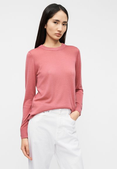 Ροζ πουλόβερ με μακριά μανίκια, κατασκευασμένο από απαλό, υφασμάτινο ύφασμα με υφή, με στρογγυλή λαιμόκοψη και διακοσμητικά κουμπιά στα μανίκια, σε συνδυασμό με λευκό παντελόνι.