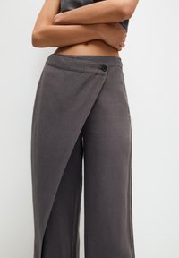 Mujer vistiendo pantalones de pierna ancha de color gris oscuro con cierre de botón, con los brazos cruzados sobre el abdomen desnudo frente a un fondo liso.