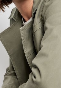 Płaszcz w oliwkowej zieleni o fakturowanej powierzchni, z usztywnionym kołnierzem i widocznymi przeszyciami. Materiał wydaje się wytrzymały i lekko pognieciony.