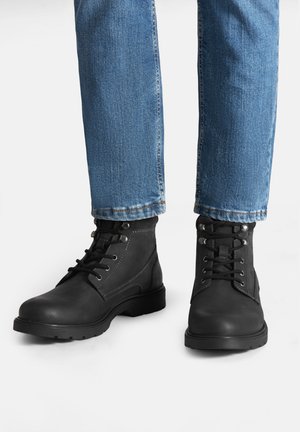 Czarne skórzane botki z okrągłymi noskami, wyposażone w metalowe przelotki, sznurowadła oraz teksturowaną podeszwę. Noszone z niebieskimi dżinsami.