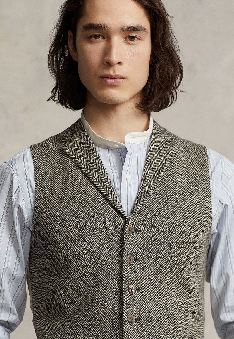 Gilet à motif pied-de-poule en tissu texturé marron et gris, avec un col cranté, une fermeture à boutons et un détail de poche avant, porté sur une chemise.