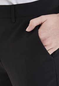 Pantaloni neri con una texture liscia; presentano una tasca frontale, un design frontale piatto e una cintura. Una mano è appoggiata nella tasca.