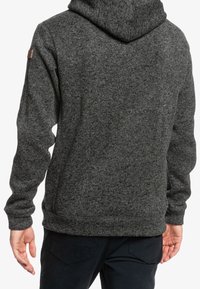 Dunkelgrauer Kapuzenpulli mit strukturierter Oberfläche, ausgestattet mit einem kleinen Logo-Patch auf der linken Schulter sowie gerippten Bündchen und Saum.