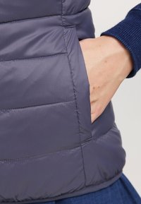 Gepolsterte Weste aus dunkelgrauem Stoff mit glatter Textur und horizontaler Steppung. Verfügt über eine Seitentasche, in der eine Hand platziert ist.