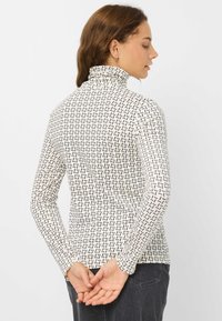 Langærmet turtleneck-top i creme med et geometrisk sort mønster, lavet af blødt stof. Har tætsiddende design og minimale sømme.