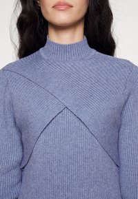 Suéter azul de punto con cuello alto, que presenta un patrón de canalé diagonal y un diseño en capas en el pecho. Textura suave.