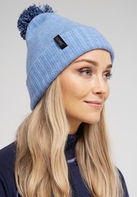 Blå strikket hue med ribbet tekstur og en marineblå pompon på toppen. Har et sort mærke på opfoldningen.