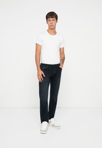 Jeans negros de corte slim, combinados con una camiseta blanca ajustada. El modelo lleva zapatillas blancas y grises, de pie frente a un fondo liso.