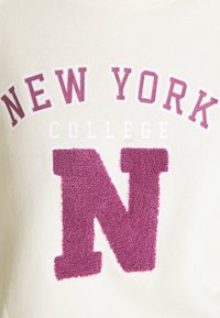 Tessuto crema con "NEW YORK" in lettere block viola, "COLLEGE" in lettere bianche più piccole e una grande "N" viola in rilievo sotto.
