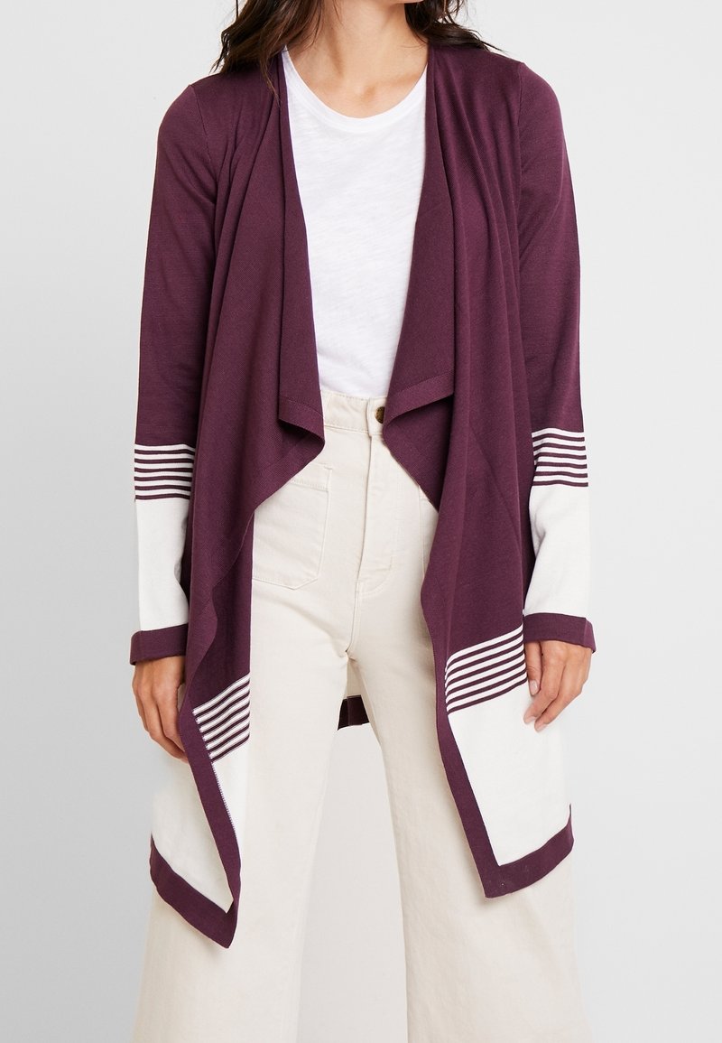 Cardigan prune à devant ouvert, accents blancs rayés sur les manches. Porté sur un t-shirt blanc et associé à un pantalon de couleur claire.