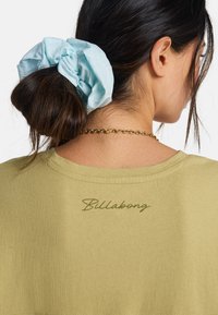 T-shirt vert olive à col rond, avec le logo "Billabong" brodé en vert près du col arrière. Chouchou en tissu bleu clair dans les cheveux.