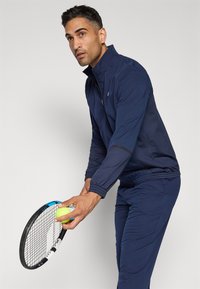 Jachetă atletică bleumarin cu guler înalt și fermoar, asortată cu pantaloni. Ține în mână o rachetă de tenis și o minge. Textura materialului este netedă.