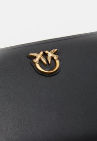Portafoglio in pelle nera con un motivo a anello di uccello in tono dorato come chiusura. La texture è liscia con dettagli raffinati sugli uccelli.
