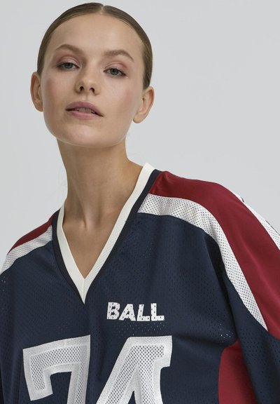 Jeune femme avec les cheveux tirés en arrière portant un maillot de sport bleu marine et bordeaux avec le numéro 74 et le mot "BALL" dessus.